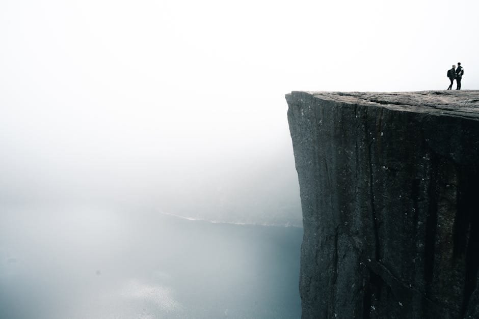 Norské fjordy | Nejúchvatnější místa v Severní Evropě 5 Two people stand on the edge of the famous Preikestolen cliff shrouded in fog, overlooking Lysefjord in Norway.