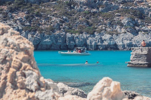 Enjoy a vibrant summer day in Rodos, Greece, with clear blue water and a boat amidst a stunning coastal landscape.