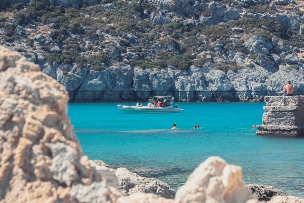 Enjoy a vibrant summer day in Rodos, Greece, with clear blue water and a boat amidst a stunning coastal landscape.
