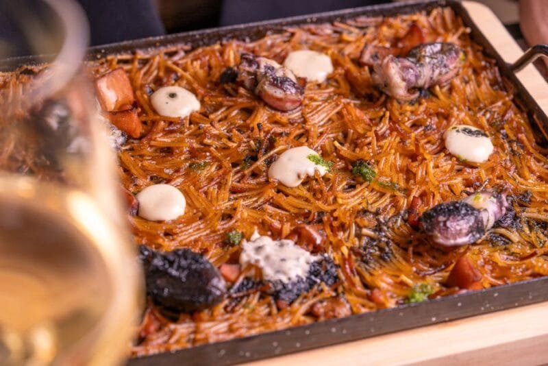 Close-up of traditional fideuà with seafood and aioli, served in a pan with a glass of wine.