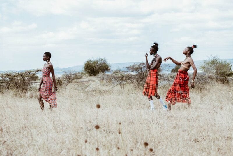 Three women in traditional attire walk across a savanna landscape, embodying cultural heritage.