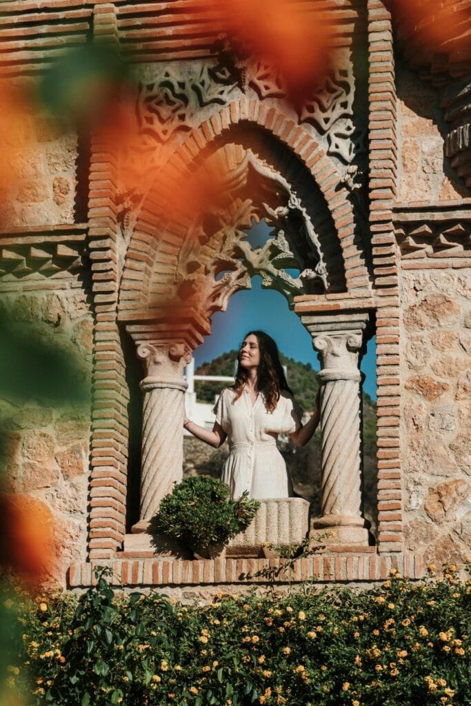 pexels photo 29151136 29151136 A woman in a light dress stands in a textured Spanish archway surrounded by vibrant flowers.